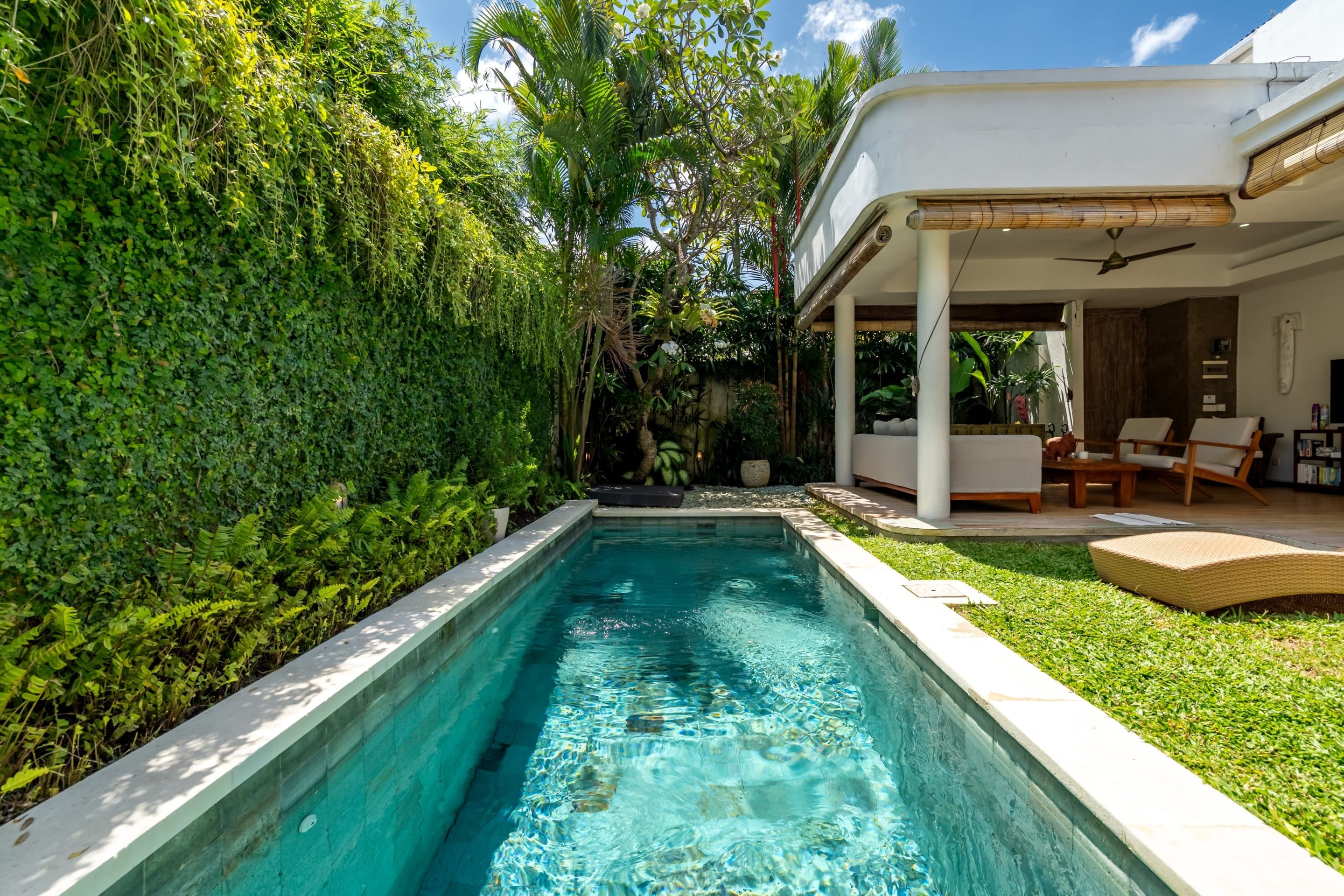 Una piscina con jardín circundante, un área de estar al aire libre cubierta y un edificio blanco con techo plano.