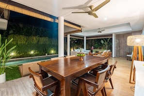Uma sala de jantar moderna com mesa e cadeiras de madeira, ventoinha no teto e vista para uma piscina e vegetação à noite.
