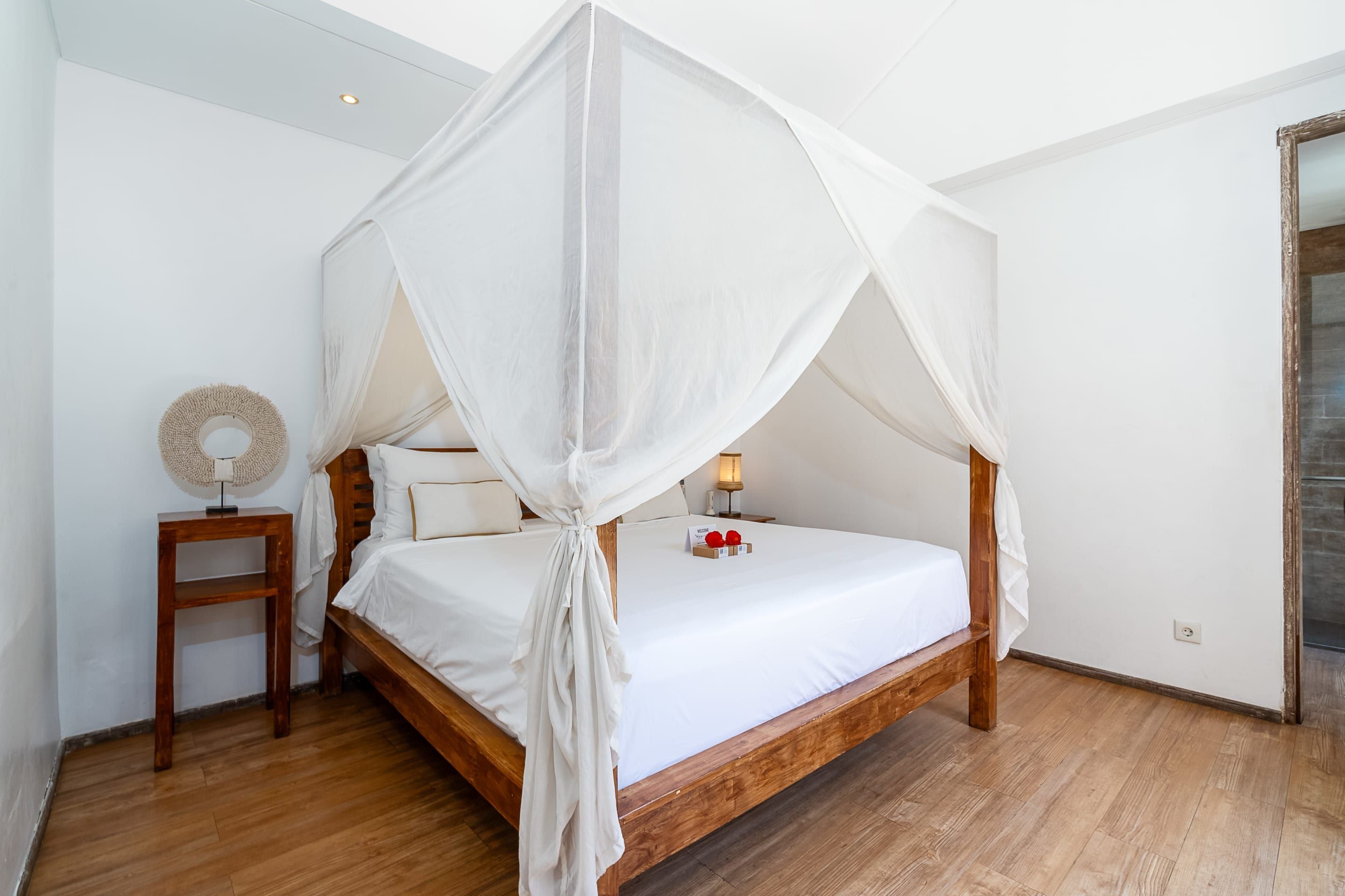A four-poster canopy bed with a wooden frame and white bedding, a bedside table with a lamp, and a wall-mounted decorative item.