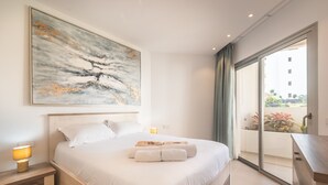 A bedroom with a large bed, a nightstand, a lamp, a painting, and a view of the outside. - Jardines - Tabor 1.2 POOL VIEW 1B (Arona)