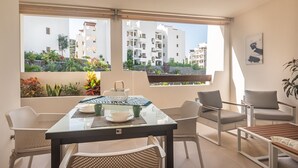 A modern living room with a glass table, wicker chairs, and a view of white buildings and greenery. - Jardines - Tabor 1.2 POOL VIEW 1B (Arona)