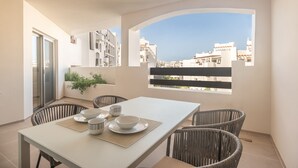 Un balcon avec une table à manger pour quatre personnes, entouré de chaises en osier, offrant une vue sur un paysage urbain avec des bâtiments blancs et un ciel dégagé.