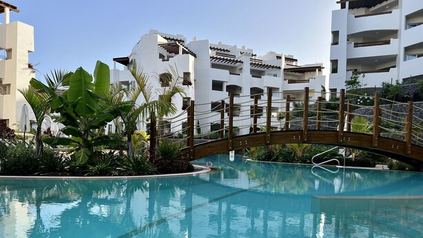 A swimming pool with a wooden bridge, surrounded by lush greenery and modern buildings. - Jardines - Adama 0.1 POOL & GARDEN VIEW 2B (Arona)