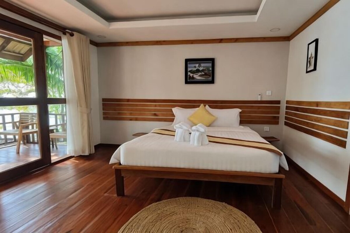 A hotel room with a large bed, wooden headboard, and a view through a glass door to an outdoor area.
