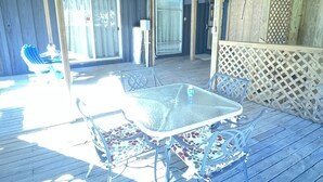 Eine überdachte Terrasse mit Glastisch und Metallstühlen, ein Holzboden und ein Gitterzaun.