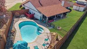 Un patio trasero residencial con piscina, un área de patio techada y un jardín cercado.