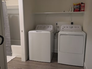 Un lavadero con dos lavadoras de carga frontal, un estante con productos de limpieza y una cortina de ducha.