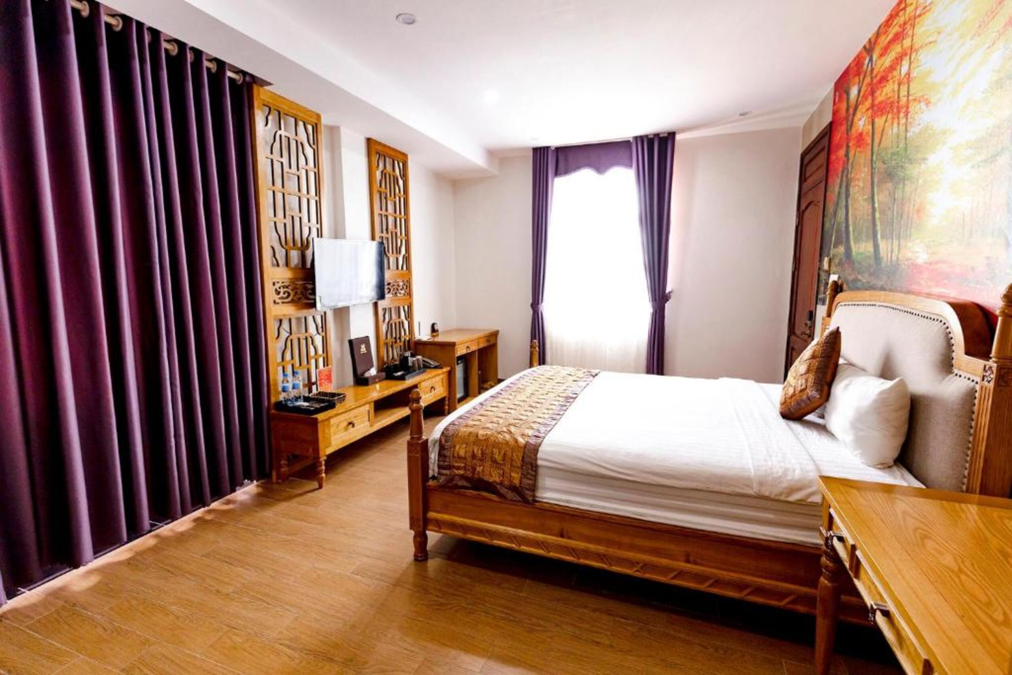 A bedroom with a wooden bed, a desk, and a chair. There is a window with curtains, a painting on the wall, and a wooden floor.