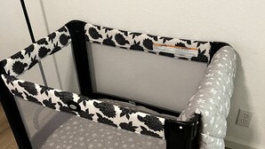 A black and white patterned crib with a gray mattress, positioned against a white wall with a framed picture above it.