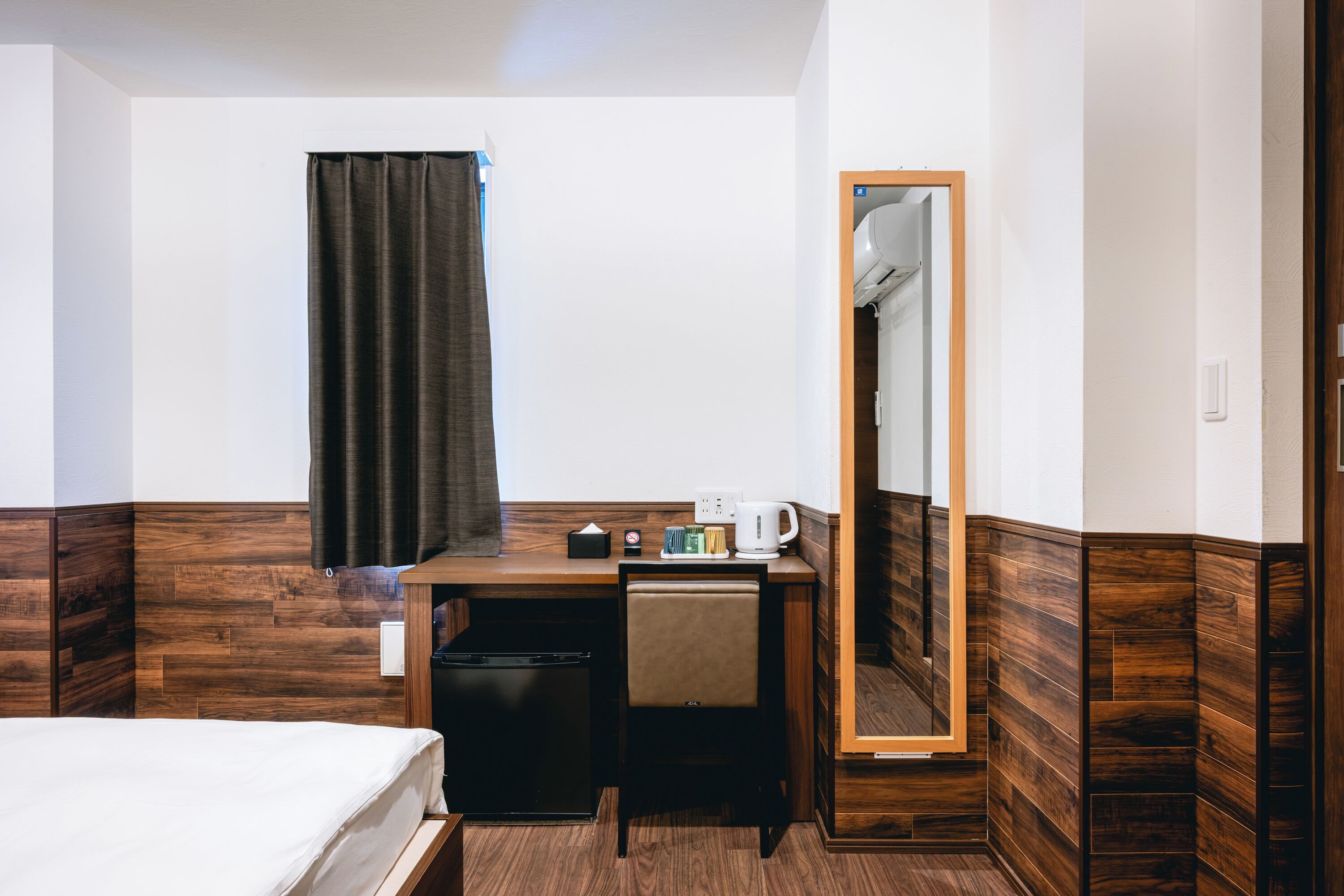 A hotel room with a bed, a desk with a chair, a mirror, and a small kitchenette.