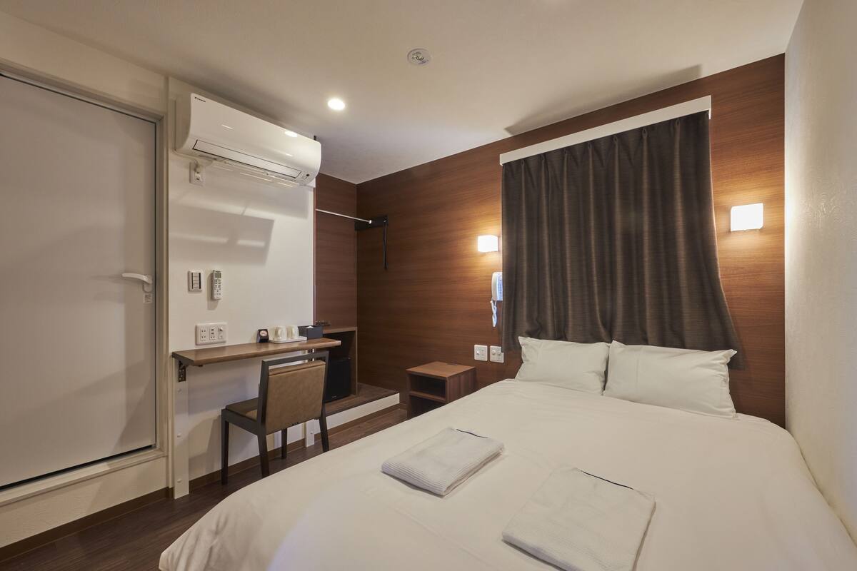 A hotel room with a bed, a desk, a chair, an air conditioning unit, and a window with curtains.