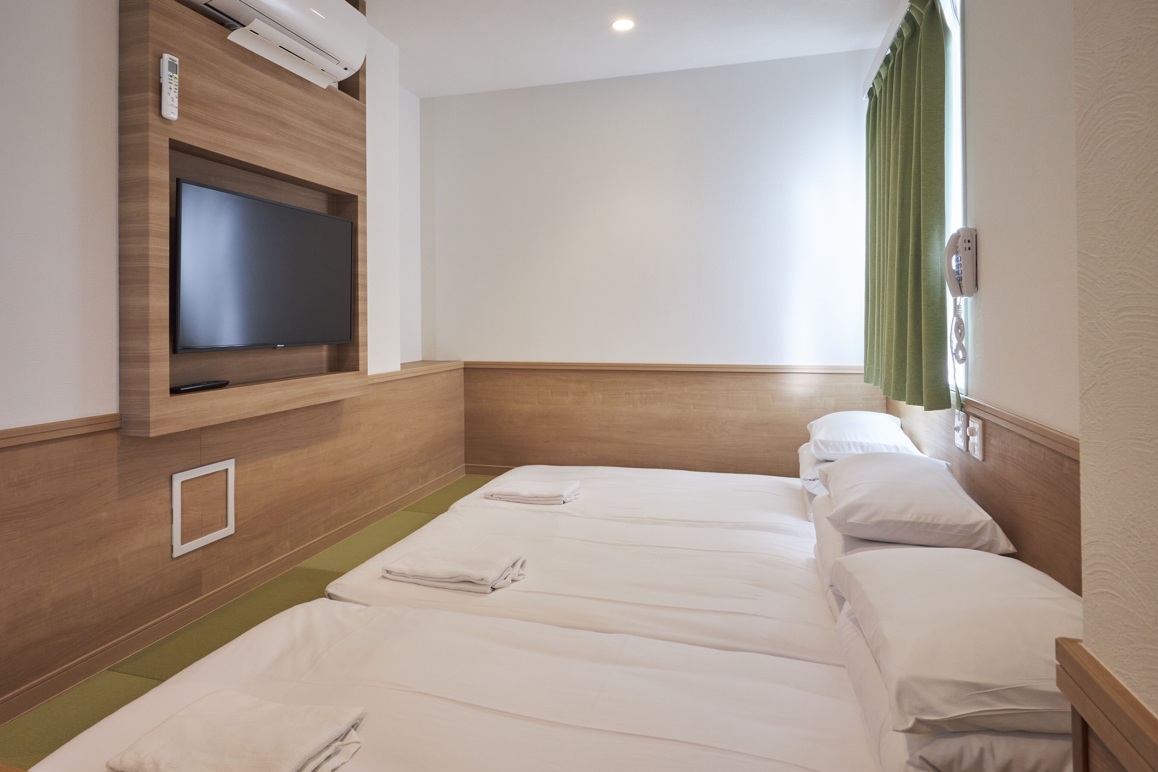 Habitación de hotel con un televisor de pantalla plana montado en una pared de madera, un ventanal con cortinas verdes y tres camas ordenadas con ropa blanca.