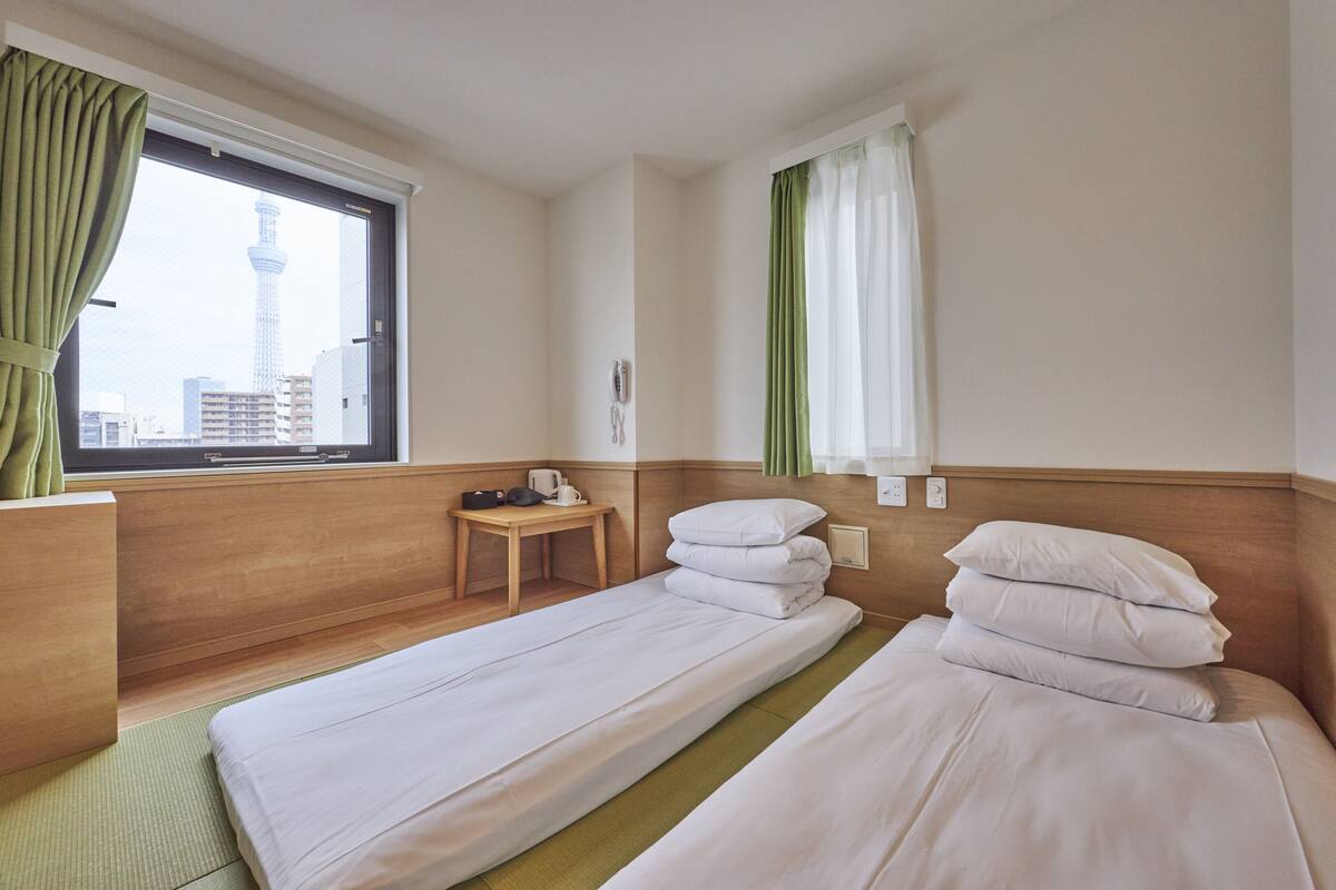 A hotel room with two beds, a small table with a kettle, and a view of a tower through the window.