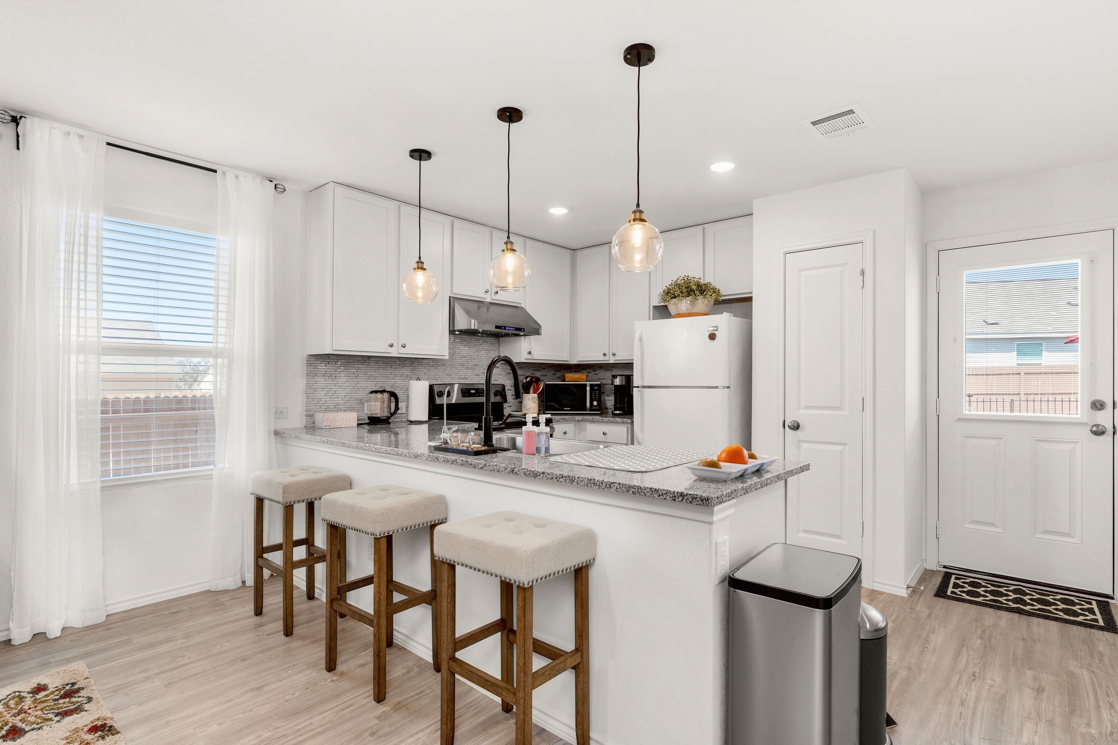 A modern kitchen with white cabinets, a stainless steel refrigerator, a gas stove, and three bar stools.