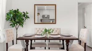 A dining room with a wooden table set for four, surrounded by upholstered chairs. A large mirror hangs above the table, reflecting a kitchen area.