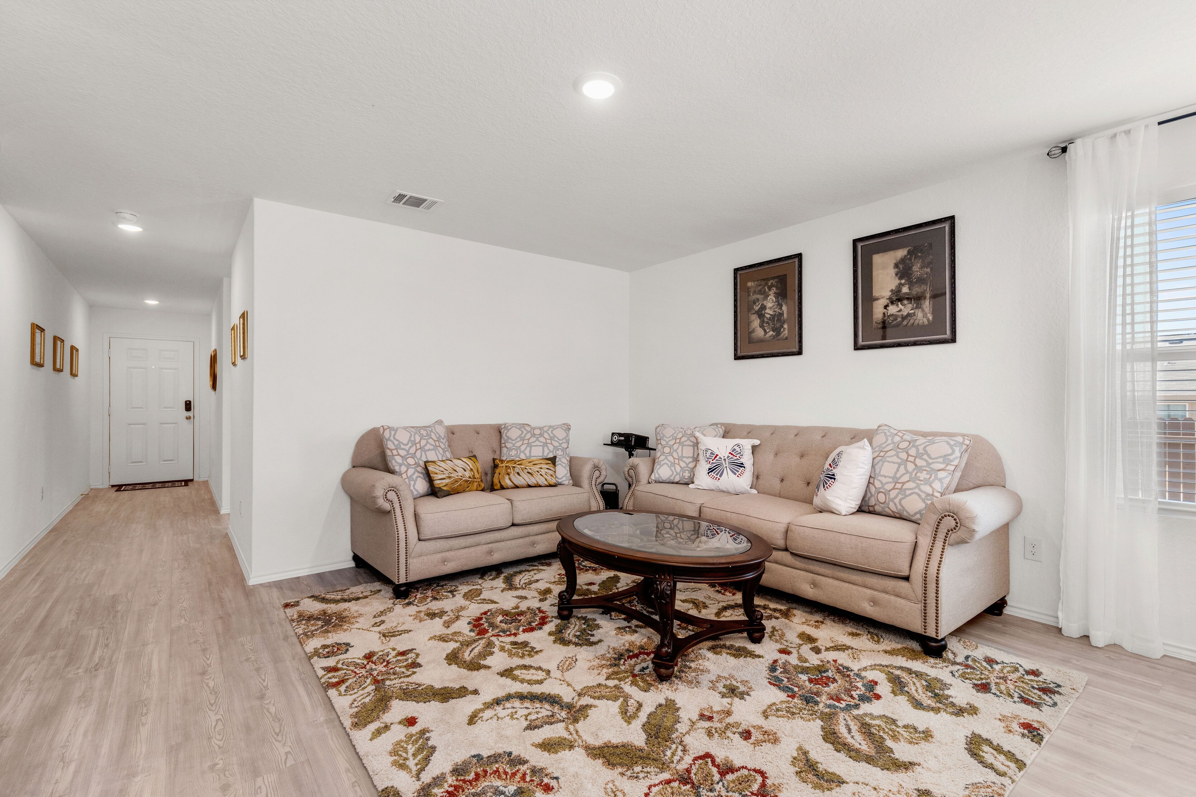 A living room with a beige sofa, a coffee table, and a patterned rug. There are two framed pictures on the wall.