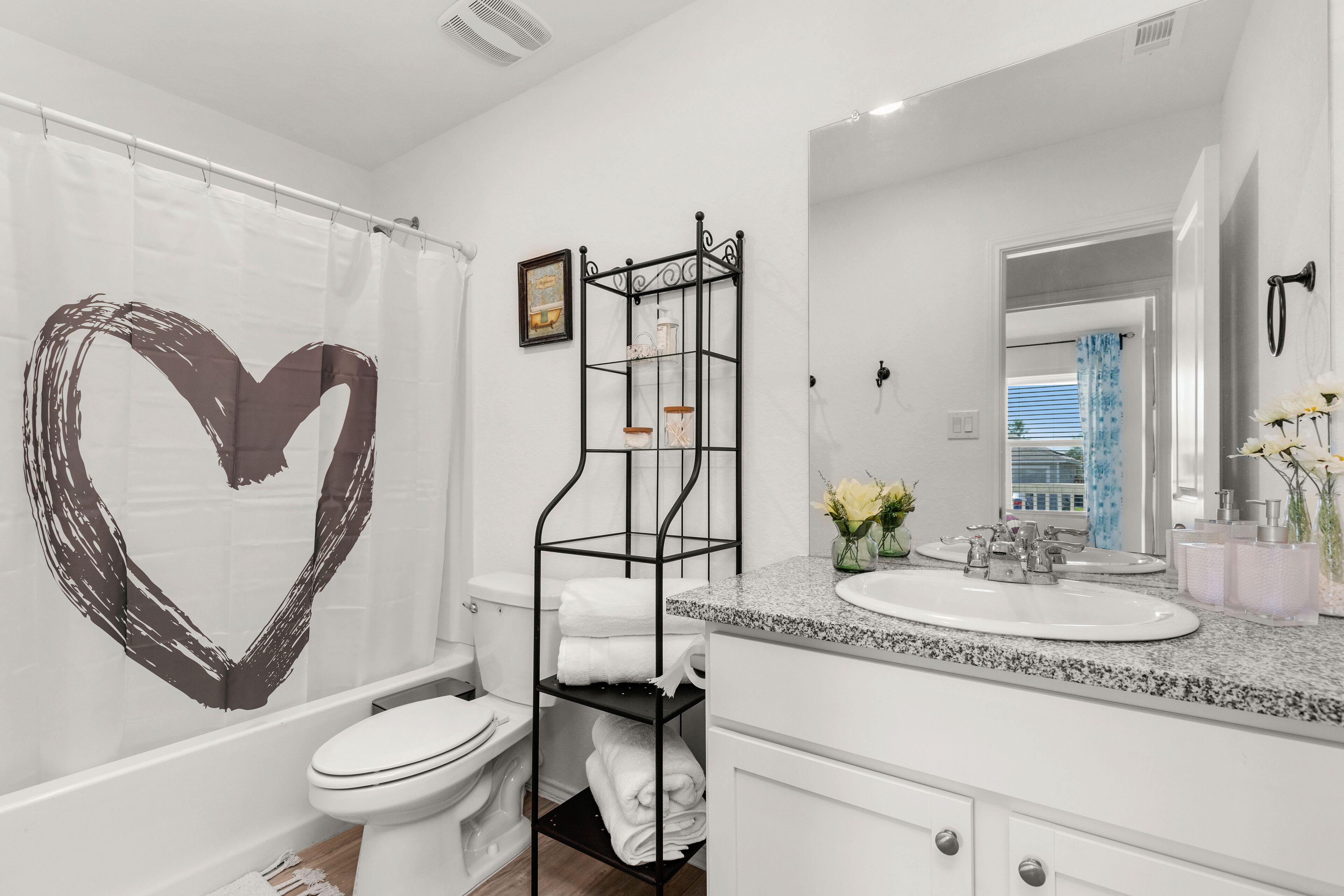 Un baño moderno con inodoro blanco, toallero negro, encimera de granito y cortina de ducha con forma de corazón.