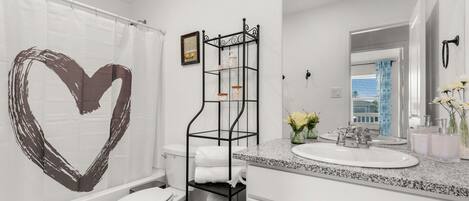Un baño moderno con inodoro blanco, toallero negro, encimera de granito y cortina de ducha con forma de corazón.