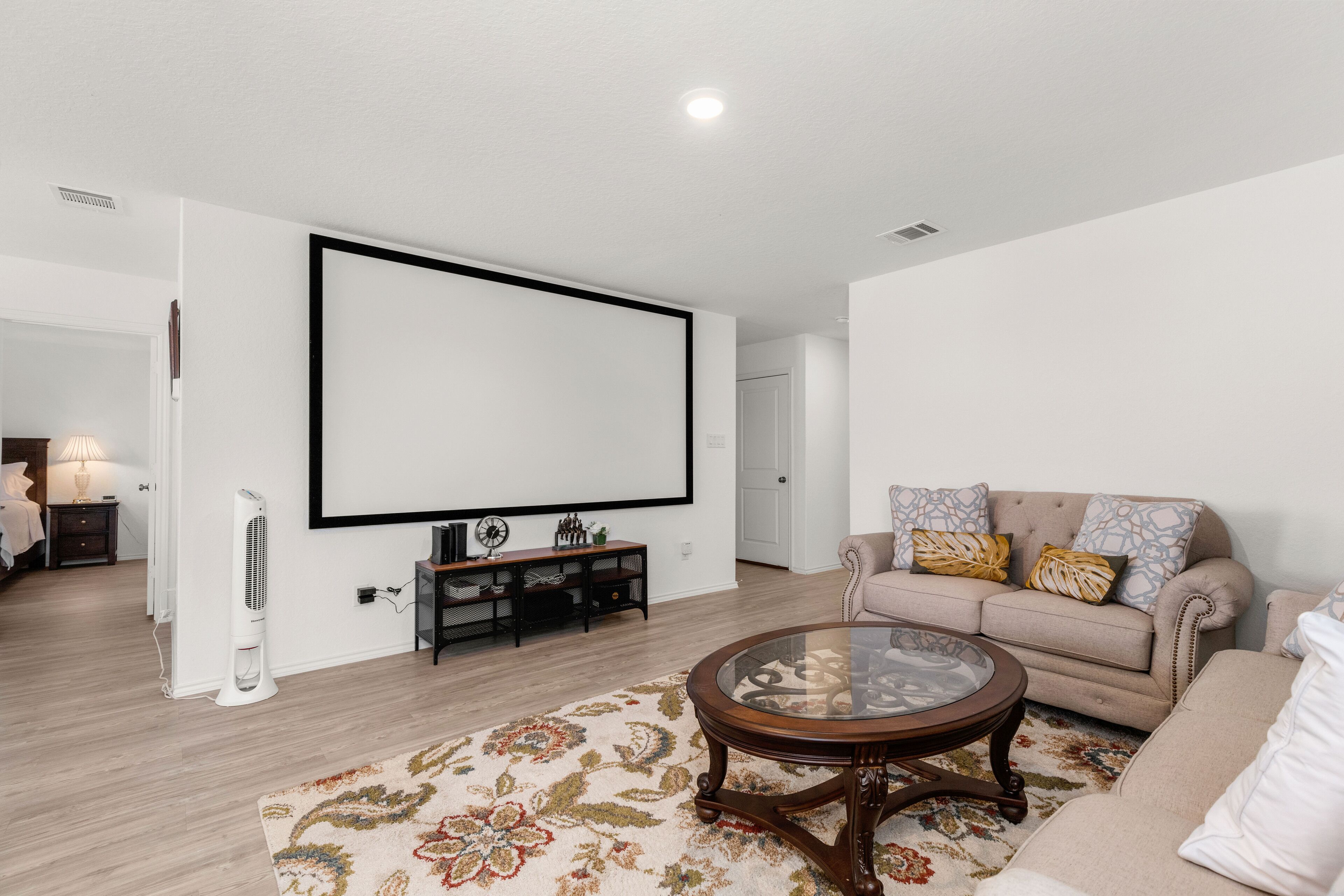 Una sala de estar moderna con un televisor de pantalla plana grande montado en la pared, un sofá color beige con cojines estampados, una mesa de centro de vidrio y una alfombra decorativa.