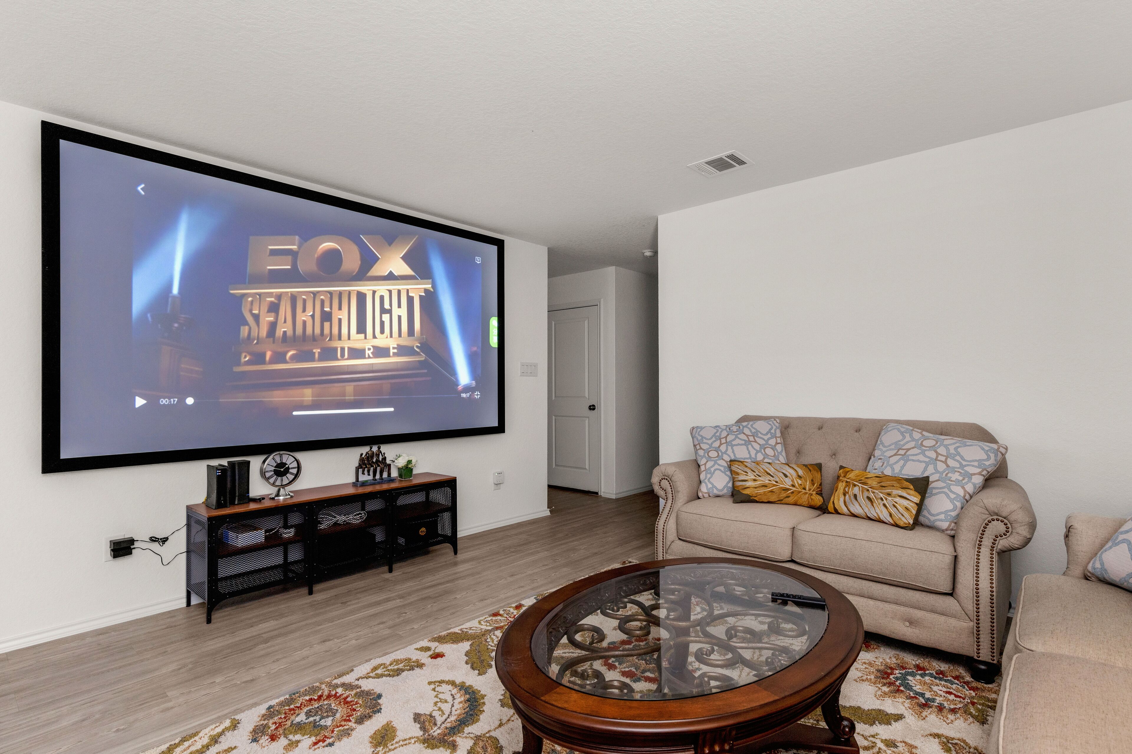 Un salón con un televisor de pantalla plana grande montado en la pared, un sofá beige con cojines estampados, una mesa de centro de madera y una alfombra con diseño.