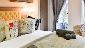Habitación de hotel con cabecera capitonada, almohadas coloridas, cama con manta texturizada y vistas al exterior a través de una ventana con cortinas.