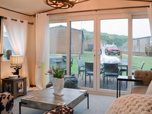 Sala de estar con sofá, mesa de centro y lámpara. Hay una puerta corrediza de vidrio que da a un patio exterior con sillas y una parrilla.