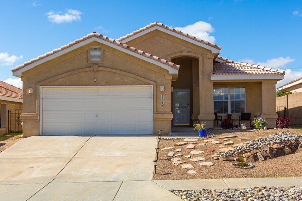 A single-story house with a white garage door, a stone pathway leading to the entrance, and a small garden with rocks and plants. - Casa Chiliwood - Short Walk to Petroglyphs / Taylor Ranch (Albuquerque)