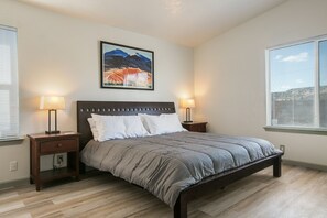 Une chambre à coucher comprenant un lit, deux tables de chevet, une commode, un tableau accroché au mur et une fenêtre avec des stores.