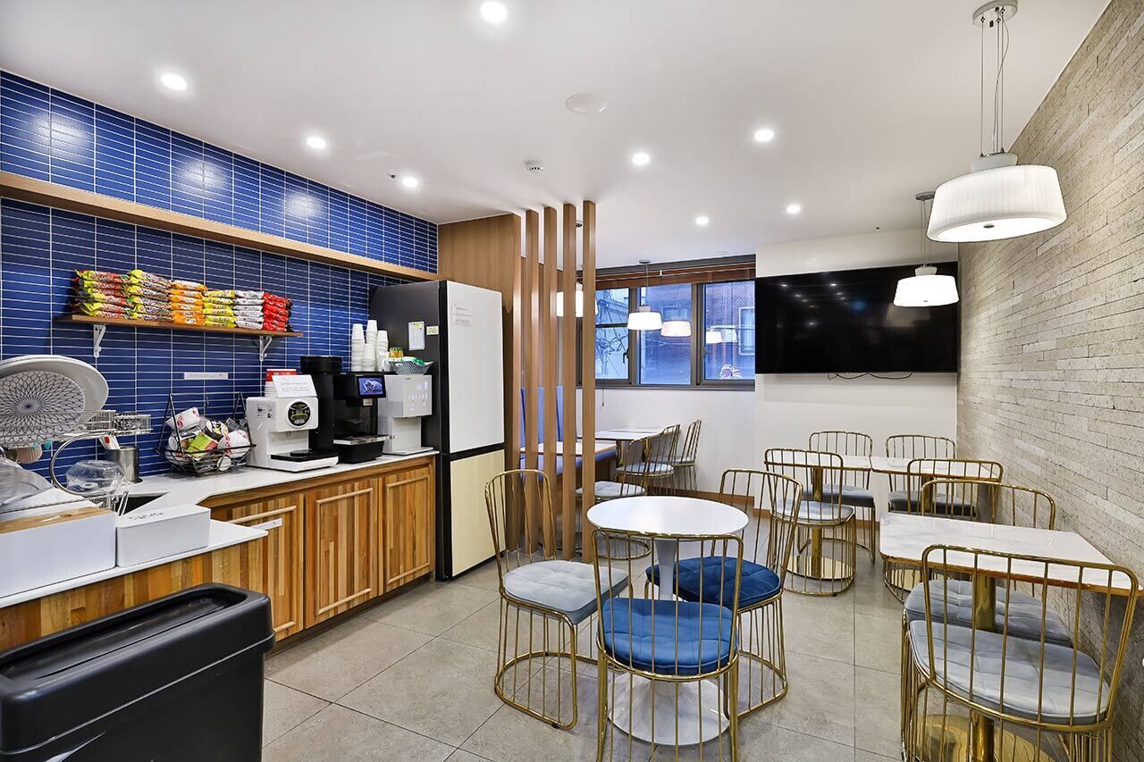 A modern kitchen with blue tiles, a coffee machine, and a counter with various items. There are round tables with metal chairs and a TV mounted on the wall.