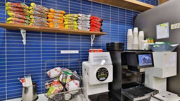 Una cafetera, una pila de fideos ramen y una estantería con alimentos envasados en un café.