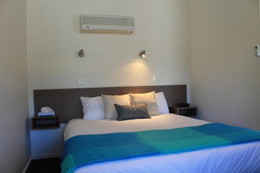 Habitación de hotel con cama, mesita de noche, aire acondicionado instalado en la pared y lámpara fijada a la pared.