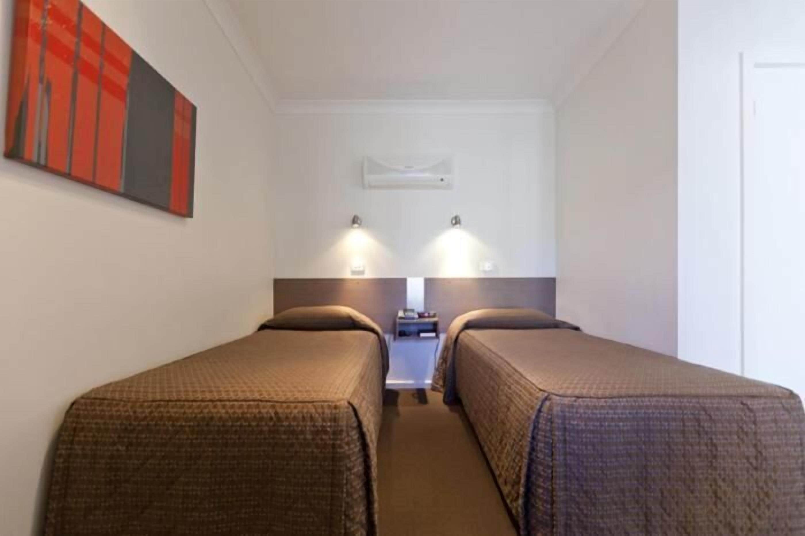 A hotel room with two single beds, a small bedside table, a nightstand, and a wall-mounted air conditioning unit.
