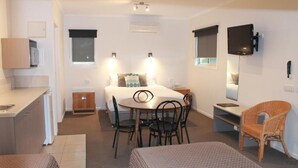 A compact hotel room with a bed, a small dining table, a sofa, a TV mounted on the wall, and a window with blinds. - Chalambar Motel (Ararat)
