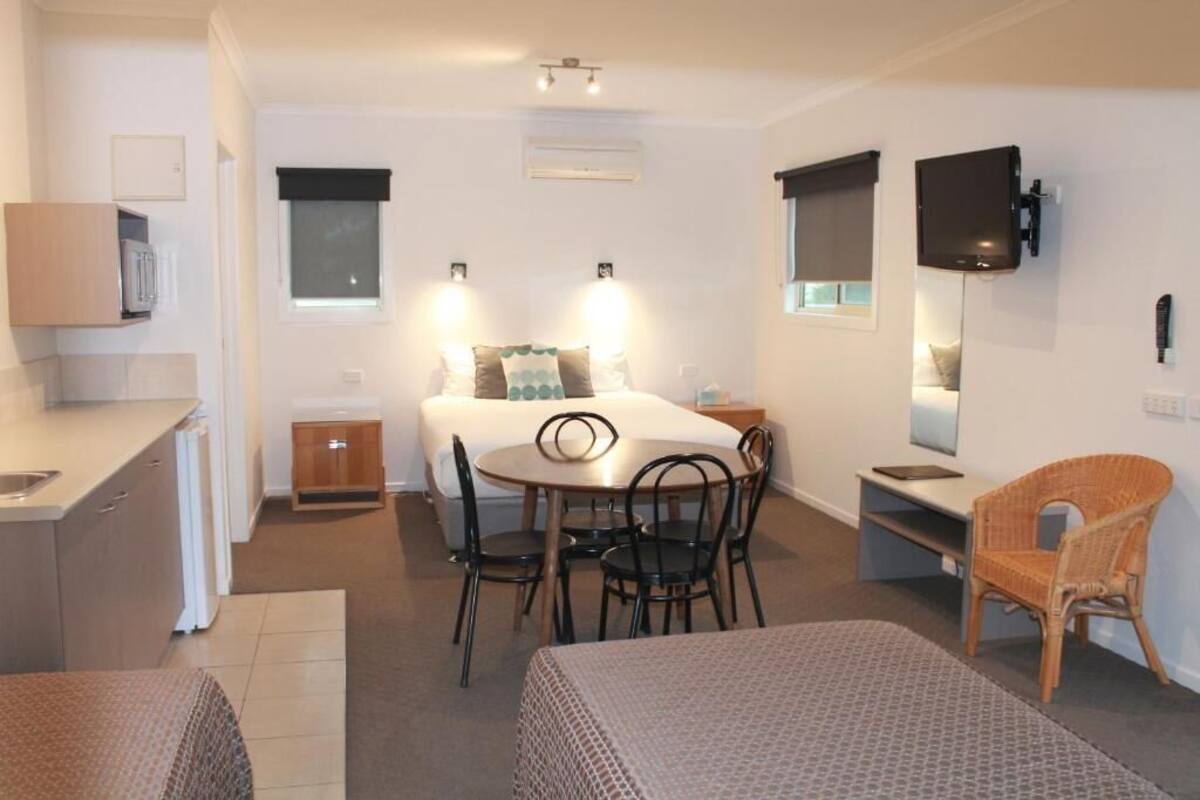 A compact hotel room with a bed, a small dining table, a sofa, a TV mounted on the wall, and a window with blinds.