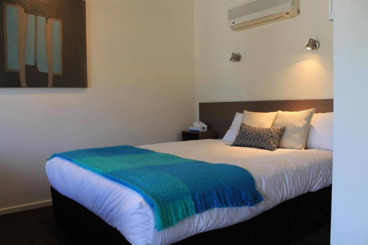 A neatly made bed with a blue and white blanket, a bedside table with a lamp, and a wall-mounted air conditioner.