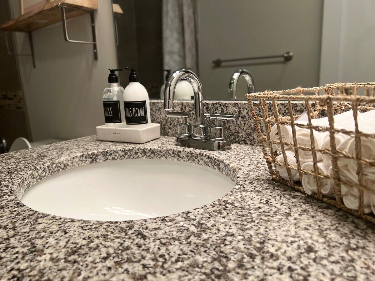Een badkamer met een granieten aanrecht, een wit wastafel en een roestvrijstalen kraan, plus een gevlochten mand met handdoeken.