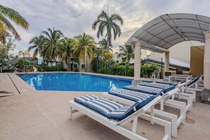 Una piscina con lettini e zona coperta, circondata da palme e edifici.