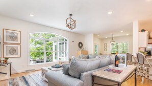 A spacious living area with a grey sofa, a coffee table, and a dining table with chairs. There are framed pictures on the wall, a chandelier, and a large window providing a view of greenery outside. - Shoreside Oasis - Amazing Views & Spacious 5BR 4BA (Mooresville)