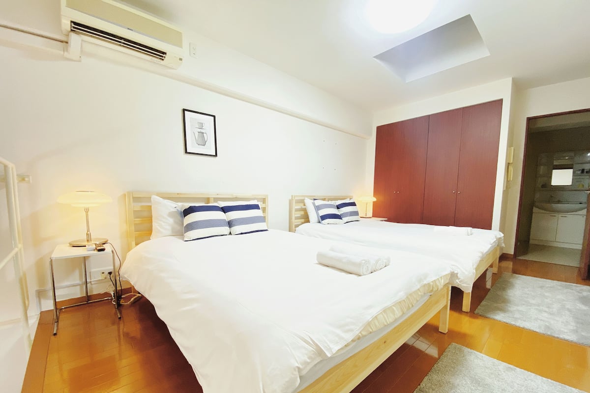 Quarto de hotel com duas camas, uma mesa de cabeceira com abajur, um quadro na parede e um guarda-roupa.