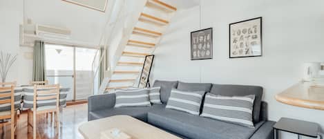 A modern living room with a grey sofa, striped pillows, a wooden coffee table, and a staircase with wooden beams.