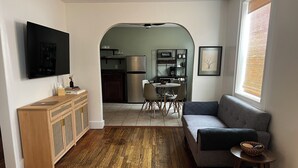 Un salon comprenant un canapé, une petite table, un téléviseur fixé au mur et une cuisine visible à travers une arche.