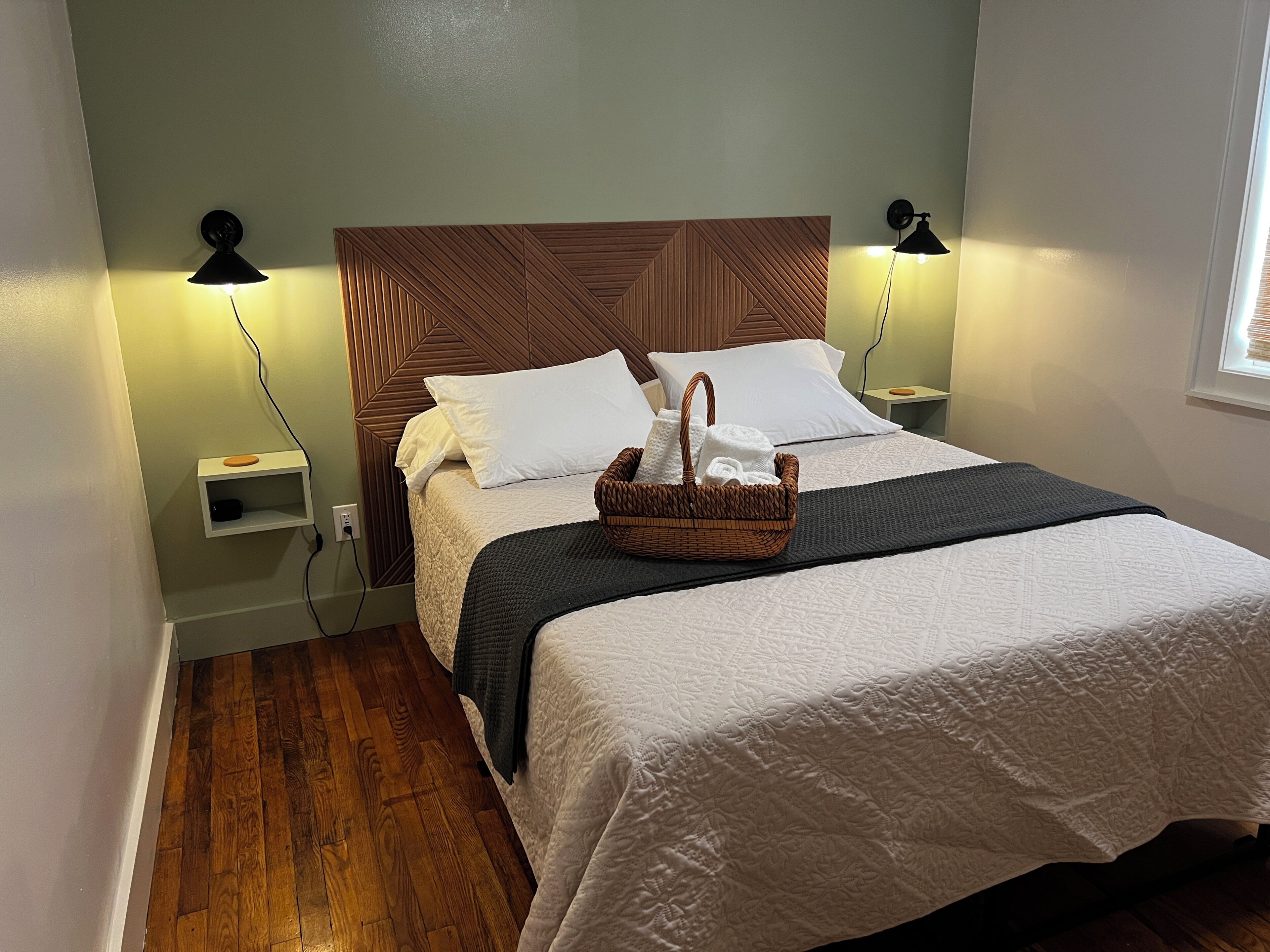 Een net opgemaakt bed met een houten hoofdbord, twee bedlampen, een klein plankje en een mandje op het bed.