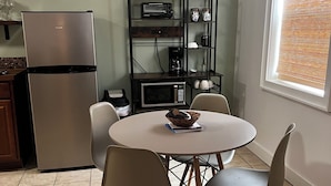 Une cuisine compacte équipée d’un réfrigérateur, d’un four à micro-ondes et d’une machine à café, le tout placé sur une étagère. On y trouve également une table à manger avec des chaises, ainsi qu’une fenêtre avec des stores.