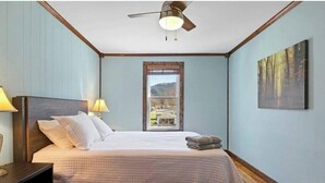 A bedroom with a bed, a nightstand, a lamp, a ceiling fan, a window with a view, and a painting on the wall.