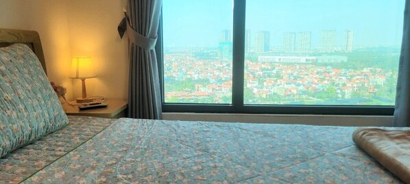 Habitación de hotel con cama, mesita de noche con lámpara y vista panorámica de la ciudad a través de amplias ventanas.