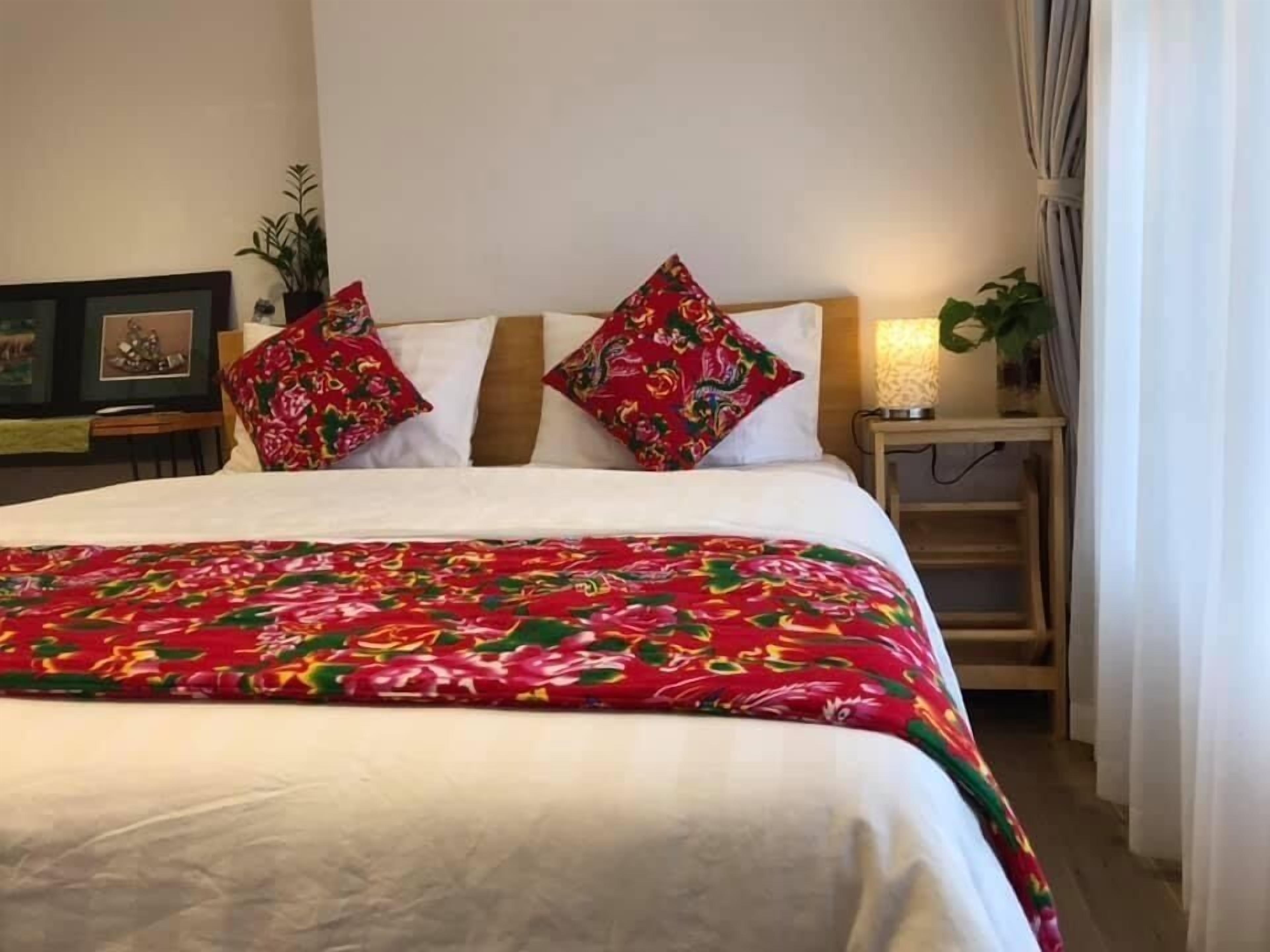 A neatly made bed with a floral bedspread, two decorative pillows, a bedside lamp, and a small nightstand with a plant.