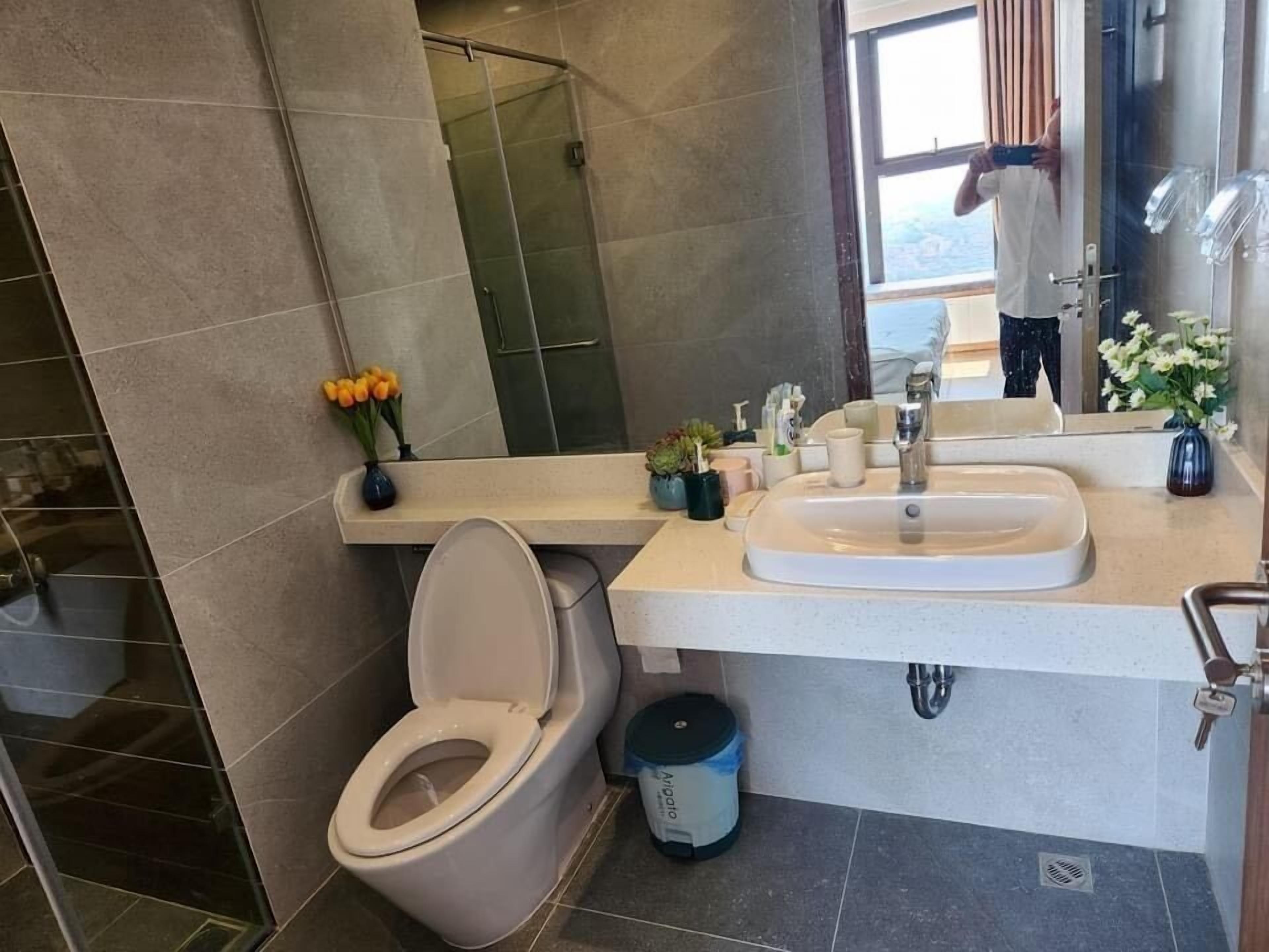 Un baño moderno con inodoro, lavamanos y ducha. Hay flores en el mostrador y un cubo de basura.