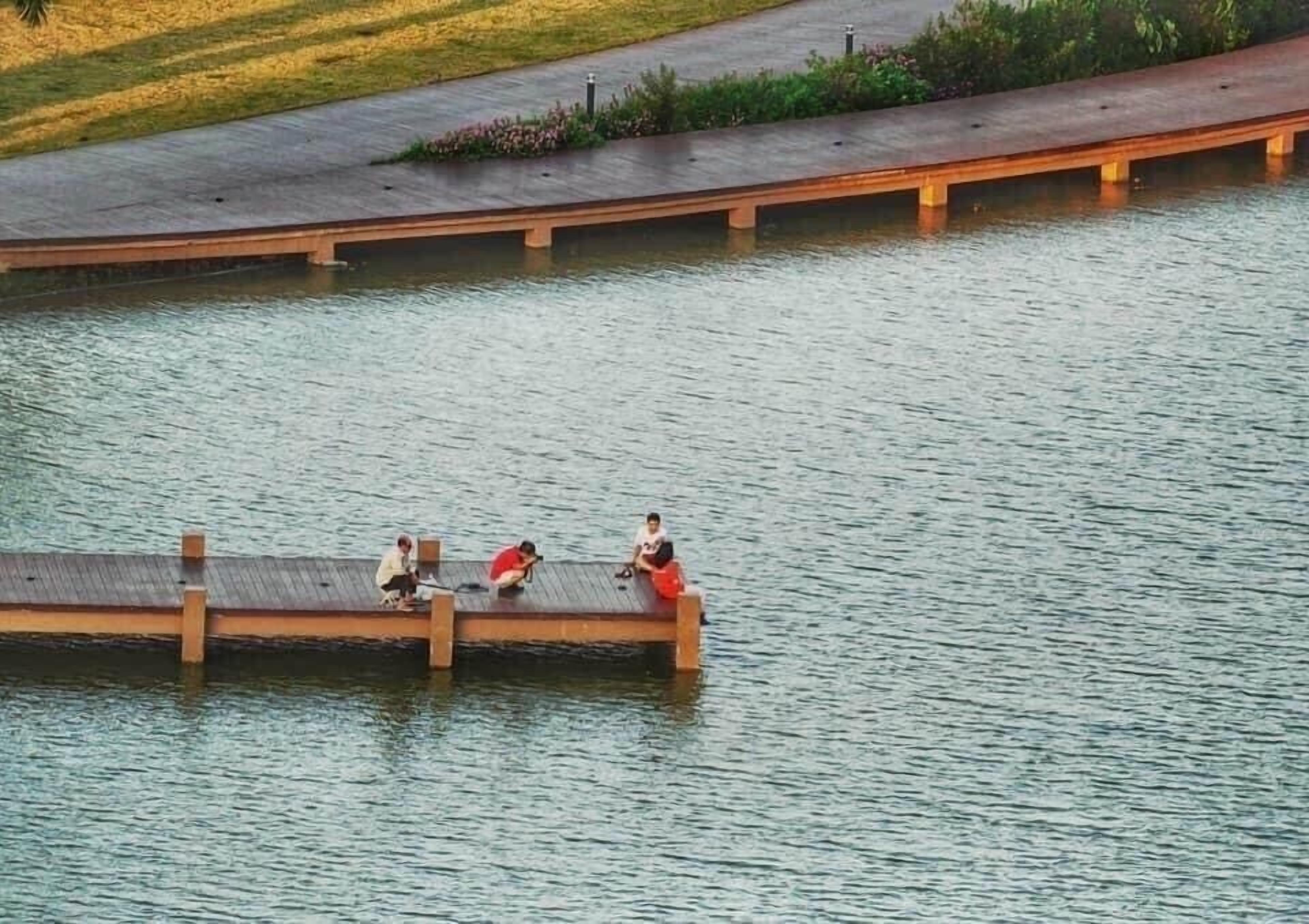 A wooden bridge over a calm body of water with three people sitting on it.