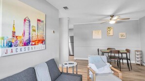 Una sala de estar moderna con un sofá gris, una zona de comedor con sillas de madera, un ventilador de techo y una obra de arte con un paisaje urbano en la pared.