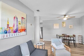 Ein modernes Wohnzimmer mit einem grauen Sofa, einem Essbereich mit Holzstühlen, einem Deckenventilator und einem Stadtbild an der Wand.